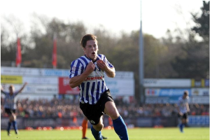 Ein Mann in einem blauen und weißen gestreiften Hemd rennt mit einem Fußball auf einem Rasenfeld während eines Spiels von Huddersfield Town gegen Swansea City in der Sky Bet Championship. Im Hintergrund sind Zuschauer, Schilder, Fahnen und Bäume unter einem klaren blauen Himmel zu sehen.