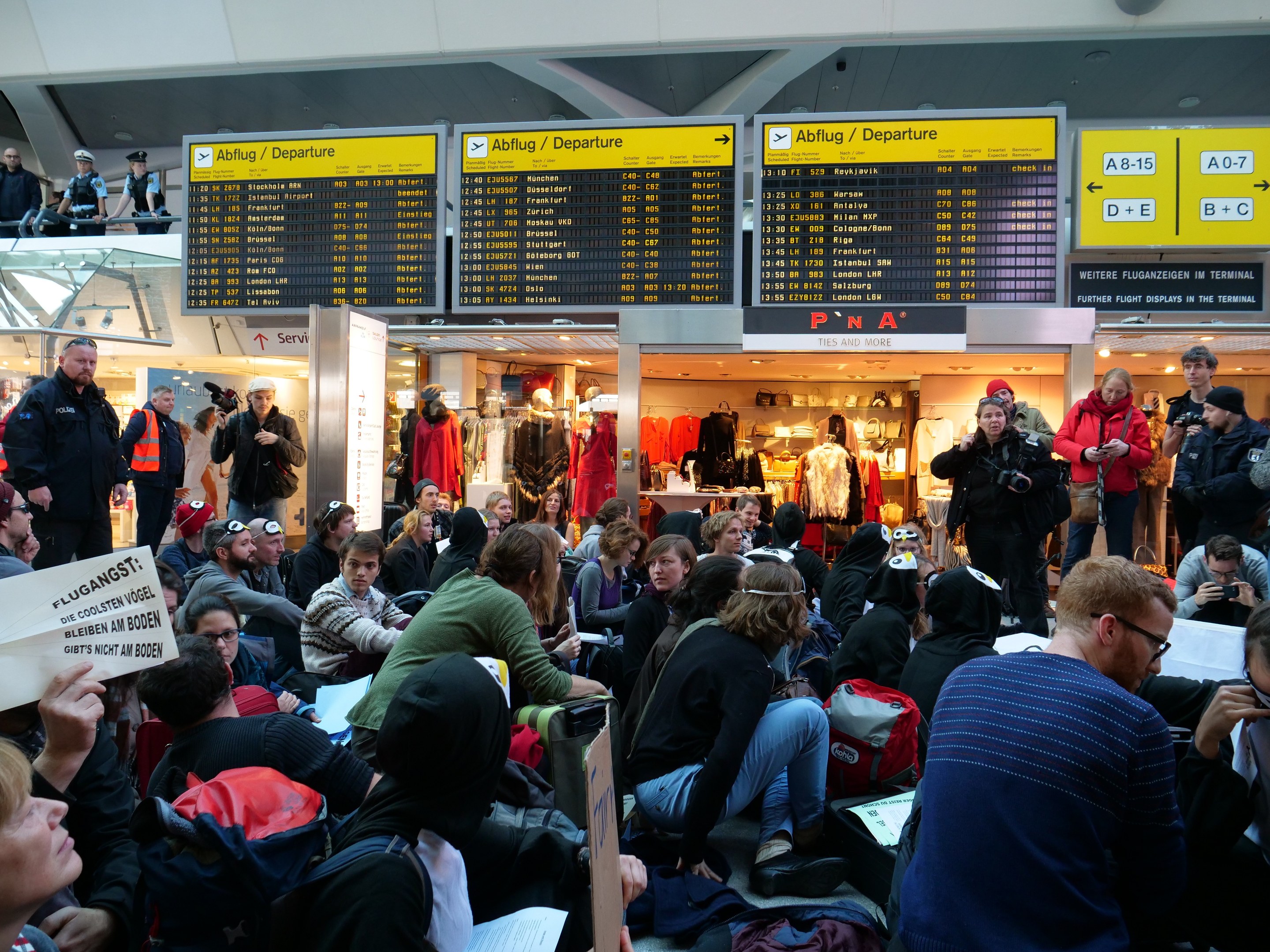 Eine große Gruppe von Menschen in einem Flughafen, einige sitzen mit Taschen und Papieren, andere stehen, mit Texttafeln, Schaufensterpuppen in Kleidern und Deckenlampen im Hintergrund, was auf eine Demonstration hinweist.