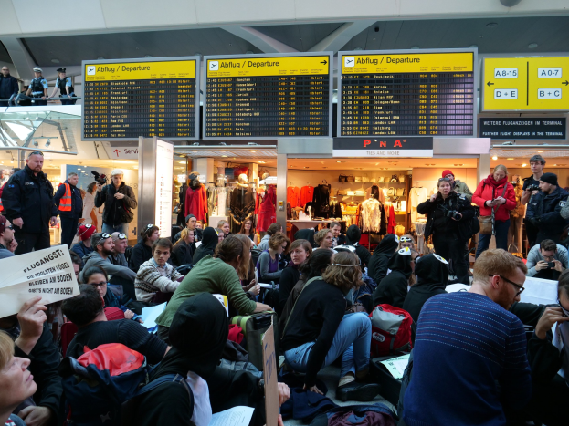 Eine große Gruppe von Menschen in einem Flughafen, einige sitzen mit Taschen und Papieren, andere stehen, mit Texttafeln, Schaufensterpuppen in Kleidern und Deckenlampen im Hintergrund, was auf eine Demonstration hinweist.