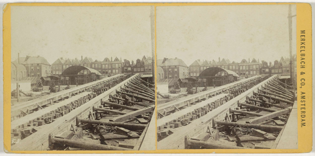 Schwarz-weiß-Foto einer Zugschiene mit Gebäuden im Hintergrund, Text 'Merkelbach & Co, Co. Amsterdam' auf der rechten Seite und hölzerne Gegenstände im Vordergrund verstreut.