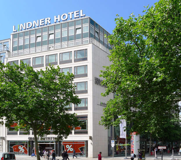 Großes Glasfrontgebäude mit "Lindner Hotel"-Text oben, umgeben von Bäumen und einer belebten Straße mit Fußgängern und Fahrzeugen unter einem sichtbaren Himmel.