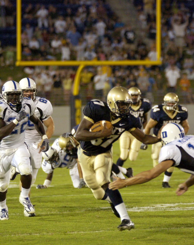 Ein Fußballspieler in Bewegung mit dem Ball, umgeben von Gegenspielern und Mannschaftskameraden in Sportkleidung und Helmen, mit einem unscharfen Stadionhintergrund und Text unten.