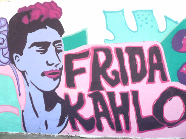 Ein Wandgemälde von Frida Kahlo an einem Gebäude in Mexiko-Stadt, das ein farbenfrohes Porträt der Künstlerin mit einer Blume im Haar und den Text 'Fridakahlo' in fetter, schwarzer Schrift zeigt.