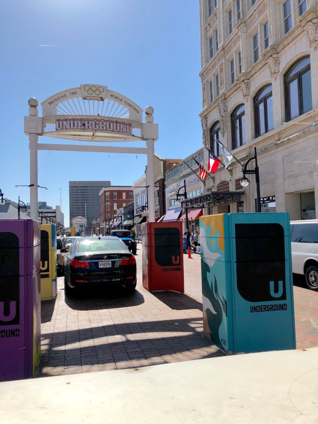 Stadtstraße mit parkenden Autos, ein Tor, Gebäude, Laternen, Fahnenmasten, ein Müllleimer und Himmel im Hintergrund.