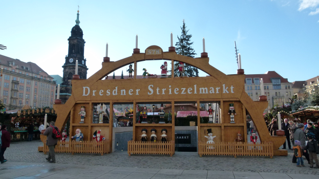 Ein geschäftiger Weihnachtsmarkt in Dresden, Deutschland, mit Menschen, die gehen, Taschen tragen und ein Tor im Hintergrund, umgeben von Bäumen, Gebäuden und einem bewölkten Himmel.