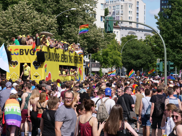 Eine große Menschenmenge marschiert auf einer Straße in Berlin, Deutschland, mit einem gelben Lastwagen im Hintergrund, viele tragen Mützen und Schutzbrillen, einige halten Fahnen, unter einem bewölkten Himmel.