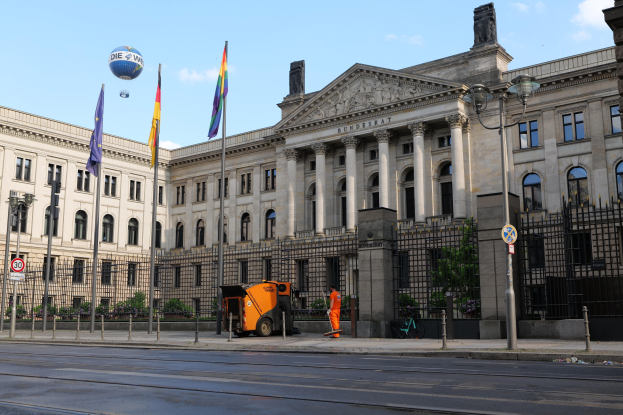 Großes Gebäude mit vielen Fenstern, als Bundestag in Berlin, Deutschland identifiziert, umgeben von Straßeninfrastruktur, Fahrzeugen, Passanten, Flaggen und Bäumen unter einem bewölktem Himmel.