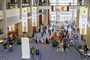 Eine große Menschenmenge bewegt sich durch ein großes Gebäude mit Bannern, Topfpflanzen, Rolltreppen, Geländern, Säulen, bedruckten Tafeln und Glastüren, einige tragen Taschen bei der Nationalen Buchmesse.