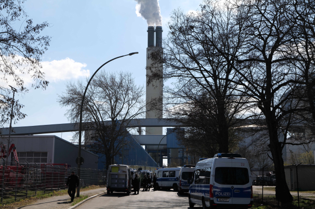 Eine Gruppe von Polizeiwagen vor einer rauchenden Fabrik geparkt, mit Menschen, Gebäuden, Bäumen und Laternen im Hintergrund unter einem bewölkten Himmel.