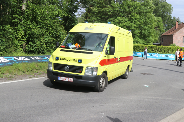 Ambulanz am Straßenrand mit einer Gruppe von Menschen in bunten Kleidern, umgeben von Bannern, Pflanzen, Bäumen, einem Haus und einem klaren blauen Himmel.