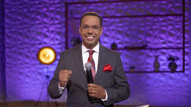 Stephen Colbert, Moderator von *The Daily Show with Trevor Noah*, steht auf einer Bühne und hält ein Mikrofon in der Hand, trägt einen Anzug, Krawatte und eine Brille, während im Hintergrund ein Scheinwerfer und ein Ständer zu sehen sind.