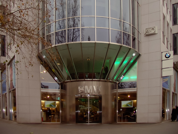 Großes Glasgebäude mit "BMW"-Text, das einen Baum auf der linken Seite und Menschen vor dem Gebäude darstellt, repräsentiert die BMW-Zentrale in Frankfurt, Deutschland.