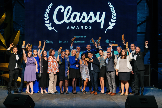 Gruppe von Menschen auf einer Bühne, die Auszeichnungen halten, mit Rednern darunter und einem Bildschirm im Hintergrund, der "Classy Awards 2019" anzeigt.