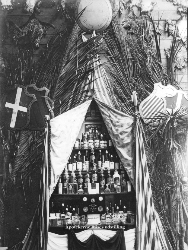 Schwarzes und weißes Foto eines Spirituosenladens mit einem Zelt in der Mitte, umgeben von Flaschen auf Regalen und Pflanzen im Hintergrund, flankiert von zwei Schilden, mit Text unten.