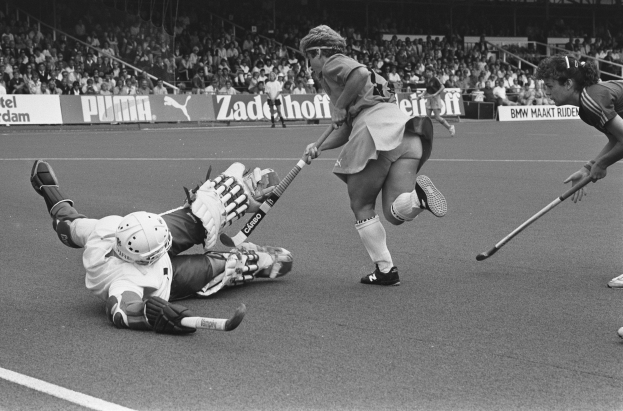 Schwarzes und weißes Bild einer Gruppe von Frauen, die auf einem Stadion Rasenhockey spielen, mit Zuschauern auf den Tribünen im Hintergrund.