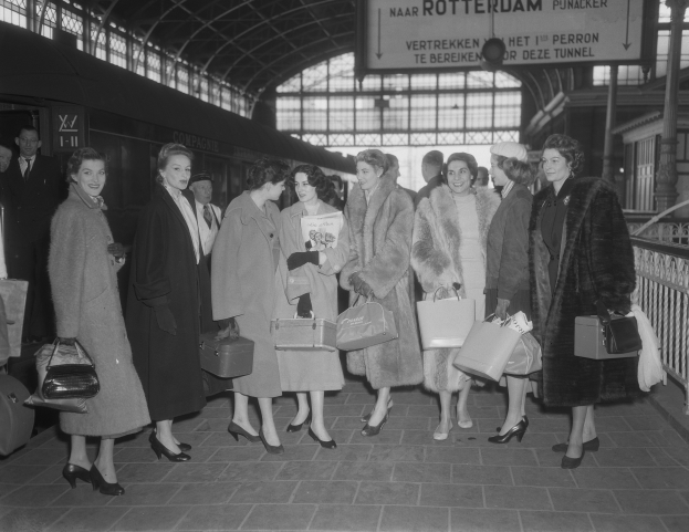 Schwarzes und weißes Bild einer Gruppe von Frauen, die an einem Bahnhof stehen, links ein Zug und rechts Geländer und eine Tafel, jede hält eine Tasche.