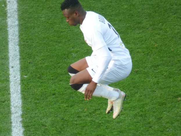 Ein Mann in weißer Uniform kniet auf einem Fußballfeld, trägt Fußballschuhe und hat eine weiße Linie auf der linken Seite des Bildes.