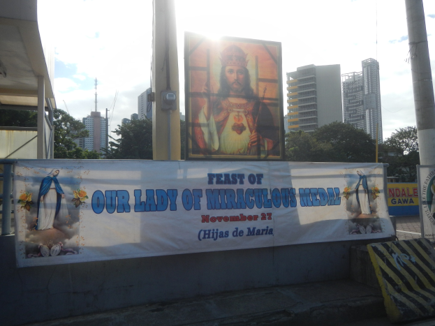 Banner mit dem Text "Fest der Unbefleckten Empfängnis" zwischen zwei Pfosten, mit einer Bank, einem Schuppen, Bäumen, Gebäuden und einem bewölktem Himmel im Hintergrund.