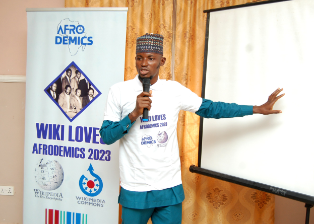Mann in einer Mütze, der ein Mikrofon hält und vor einer Whiteboard, einem Banner, einem Vorhang und einer Schalttafel präsentiert, mit einem Fokus auf 'Wiki-Liebe der Afrodemics.'