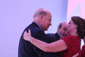 Ein Mann und eine Frau umarmen sich herzlich auf einer Bühne, umgeben von anderen, wobei der Mann ein Armband trägt.