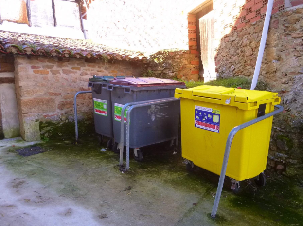 Zwei gelbe und graue Recyclingcontainer mit Rädern stehen nebeneinander vor einem Gebäude mit einer Wand und einem Fenster im Hintergrund.