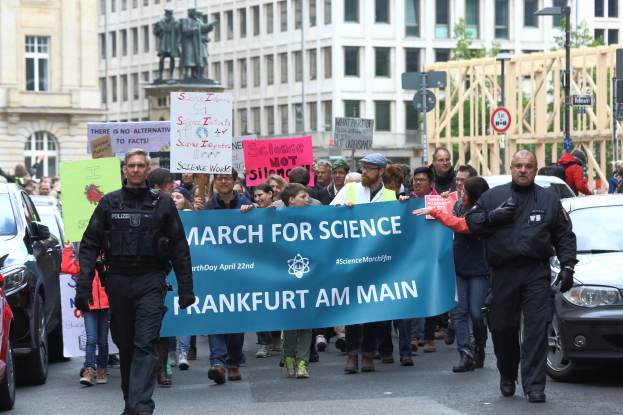 Eine Gruppe von Menschen marschiert auf einer Straße und hält ein «March for Science Frankfurt am Main»-Schild, mit Autos, Gebäuden, Statuen, Laternenmasten, Schildern und Bäumen im Hintergrund.