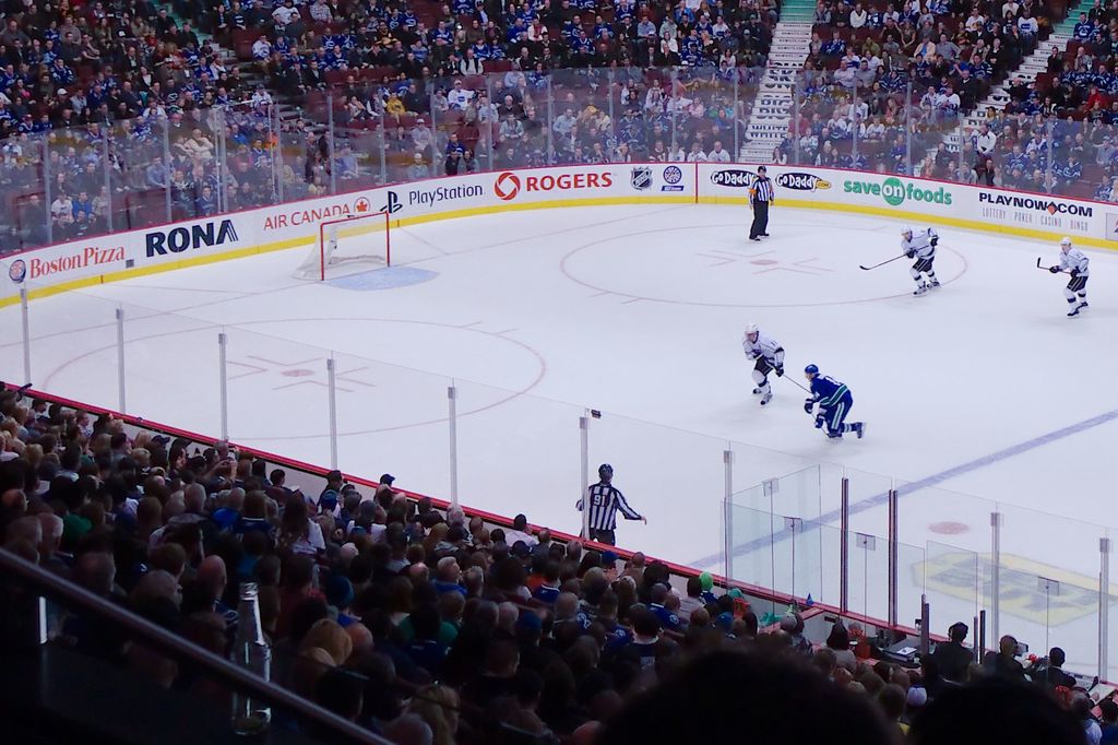 Vier Männer spielen Hockey auf einem Eisstadion, mit zwei in der Nähe des Tors und Zauns stehend, Zuschauern auf Stühlen im Vorder- und Hintergrund sowie einer Treppe im Hintergrund.