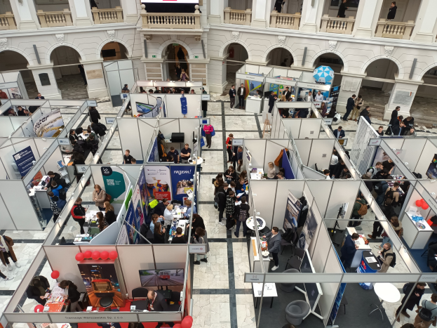 Große Gruppe von Menschen auf einer internationalen Fachmesse mit Ständen, Bannern, Tischen, Stühlen und Ballons in einem Raum mit architektonischen Bögen, Säulen, Geländern und einem Bildschirm im Hintergrund.