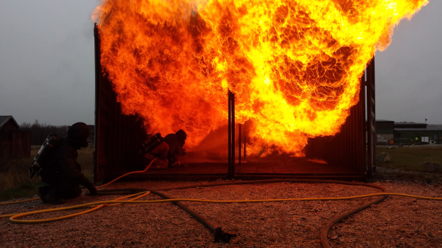 Feuerwehrleute löschen ein Gebäude Feuer mit einem auf dem Boden liegenden Schlauch, umgeben von Schuppen, Bäumen und einem klaren blauen Himmel.