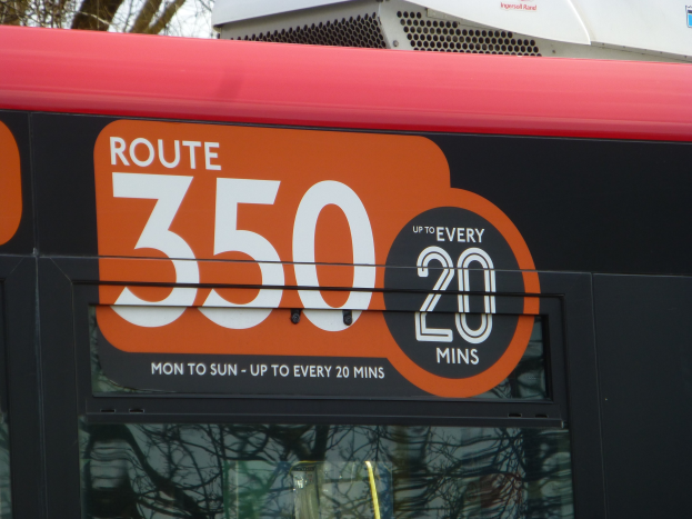 Roter Doppeldeckerbus mit der Aufschrift "Route 350" fährt durch einen baumbewachsenen Bereich mit sichtbarem Himmel im Hintergrund.
