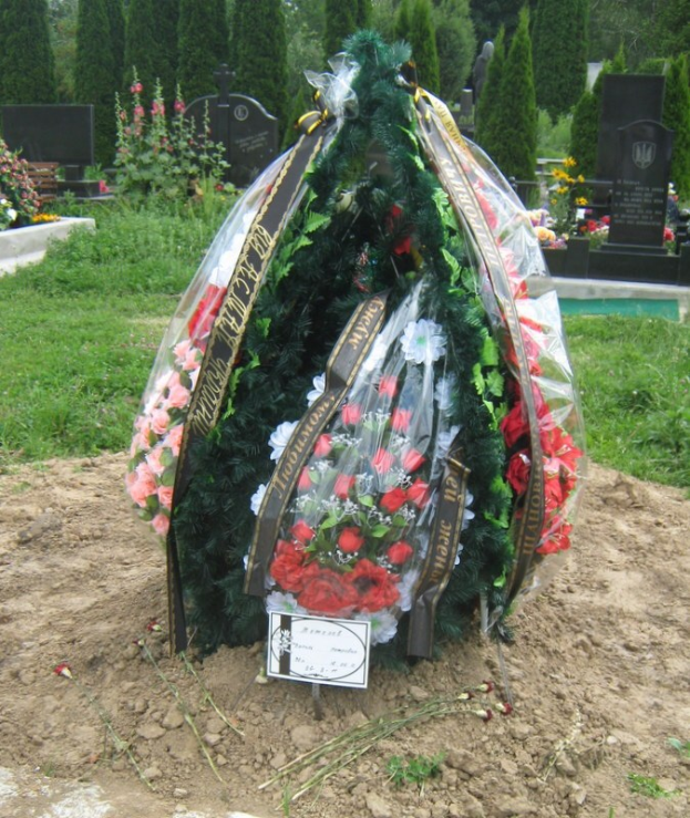 Ein Sarg mit einem farbenfrohen Blumenkranz darauf, der auf einem Friedhof umgeben von Grabsteinen, Bäumen und einem klaren blauen Himmel steht.