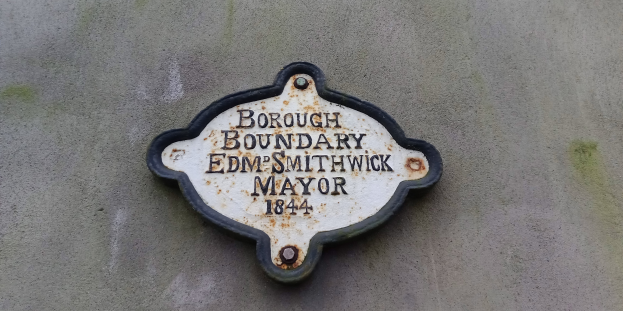 Metallplatte an der Seite eines Gebäudes mit schwarzem Hintergrund und weißer fettgedruckter Schrift, die "Borough Boundary Edm Smithwick Mayor" liest.