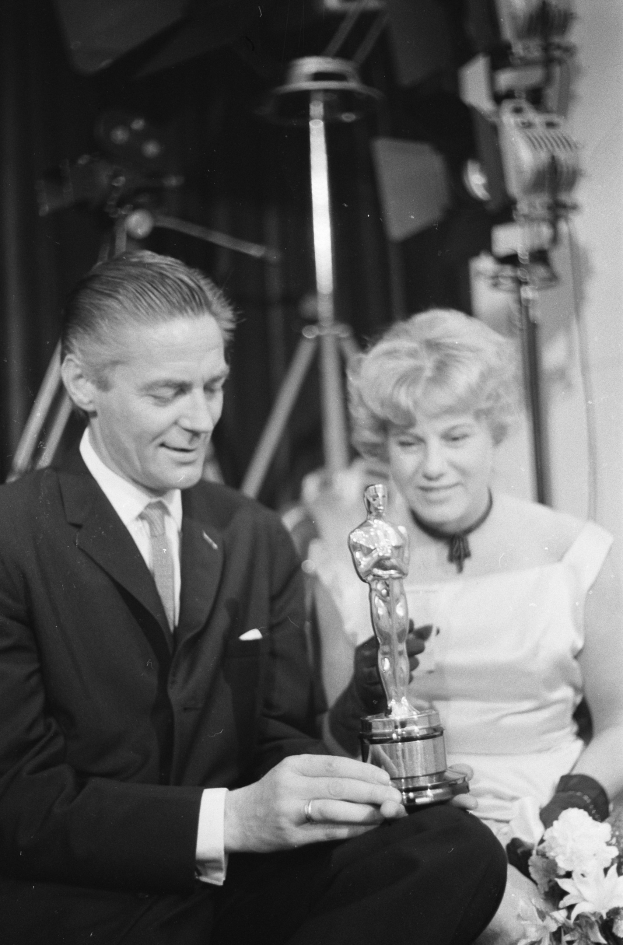 Ein Mann in einem schwarzen Blazer, weißem Hemd und Krawatte hält eine Oscar-Statue in der Hand und sitzt neben einer Frau, die einen Blumenstrauß hält, mit Scheinwerfern im Hintergrund, alles in Schwarz-Weiß.