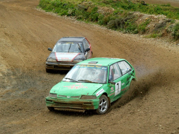 Zwei Autos mit Text und Zahlen fahren auf einer Schotterstraße durch Gras, Pflanzen und Steine, was auf ein Rallye hinweist.