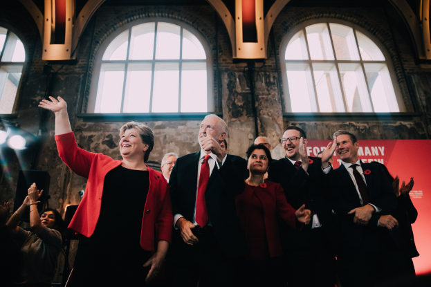 Eine Gruppe von Menschen, Mitglieder der Labour Party, klatschen feierlich vor einer Menge, mit einem Banner und beleuchteten Fenstern im Hintergrund.