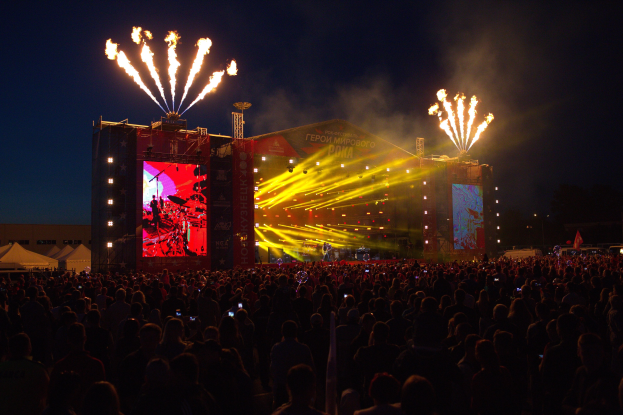 Eine große Menschenmenge auf einem Musikfestival schaut auf einer hell beleuchteten Bühne Performern zu, mit Zelten links und Feuerwerk am dunklen Himmel dahinter.