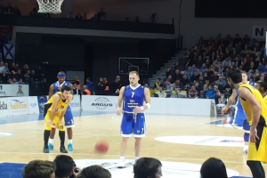 Eine Gruppe von Männern in blauen und gelben Uniformen spielt Basketball auf einem Court während des 7days Eurocup Regular Season Round 2 Spiels zwischen Fenerbahce und Real Madrid, mit Zuschauern auf den Tribünen und einem Basketballkorb am oberen Ende des Courts.