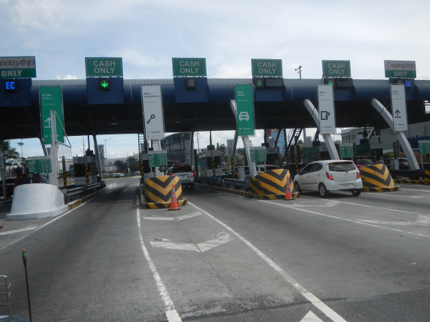 Eine Mautstation an der Seite einer Stra√e mit darunterfahrenden Autos, umgeben von Verkehrskegeln, Absperrpfölen, Schildern, einem Tor, Laternenmästen, Bäumen, Gebäuden und einem bewölktem Himmel im Hintergrund.