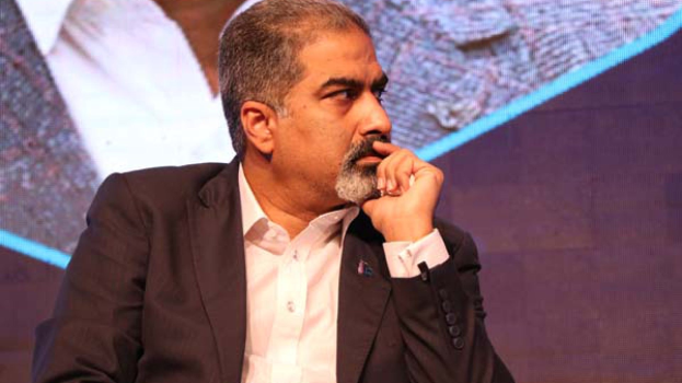 Ein Mann im Anzug sitzt auf einer Bühne und denkt nach, mit einem Bildschirm im Hintergrund.