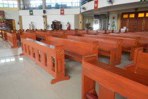 Das Innere einer Kirche mit hölzernen Kirchenbänken in Reihen, einem gefliesten Boden, einer verzierten Wand mit Fotorahmen und anderen Gegenständen, einer Decke mit Lüftern und Glasfenstern.