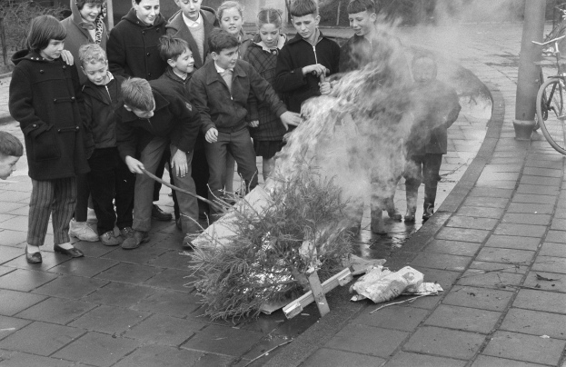 Schwarz-weißes Bild von Kindern, die sich um ein Straßenfeuer scharen, einige halten Stöcke, mit Fahrrädern, Pfählen, Bäumen, Pflanzen und Gebäuden im Hintergrund.