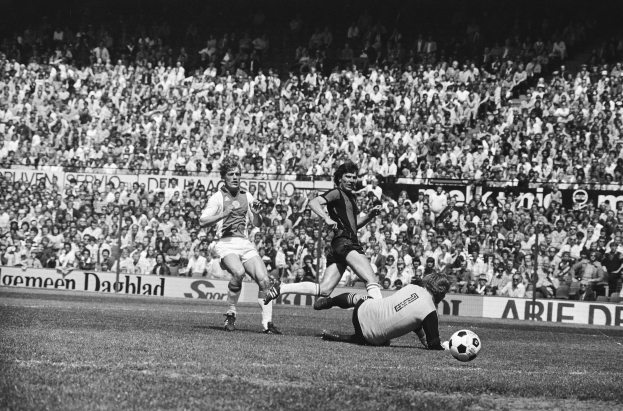 Schwarzes und weißes Foto von Männern, die Fußball spielen, auf einem Feld mit Zuschauern im Hintergrund und einer 'Bundesliga 1978-79 Ajax 1-1 Hannover 96' Flagge.