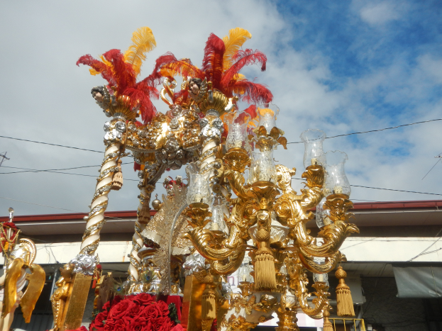 Ein großes goldenes und rotes Festwagen, das mit Blumen und anderen Dekorationen geschmückt ist, in einem Karnevalsumzug, mit einem Gebäude, Strommasten mit Drähten und einem bewölkten Himmel im Hintergrund.