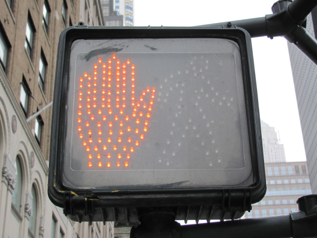 Eine Ampel mit einer roten Hand in der Mitte einer Stadt, umgeben von Gebäuden auf beiden Seiten und einem klaren Himmel darüber.