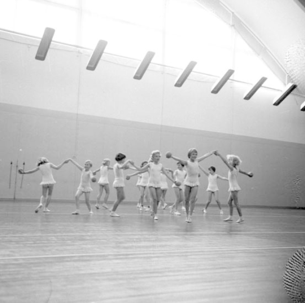 Schwarz-weißes Bild von jungen Mädchen, die in einem Studio tanzen und sich an den Händen halten, mit einem Ball auf der rechten Seite und Lampen an der Decke; ein wandmontiertes Objekt ist im Hintergrund sichtbar.
