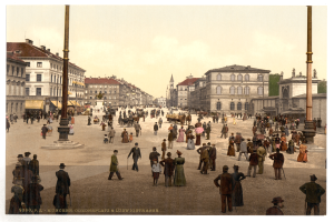 Ein altes Foto einer belebten Münchner Straßen Szenerie aus den frühen 1900er Jahren, das Fußgänger, einige mit Schirmen, einen Pferdewagen im Vordergrund, Gebäude, Bäume und Pfosten im Hintergrund zeigt, mit Text am unteren Rand des Bildes.