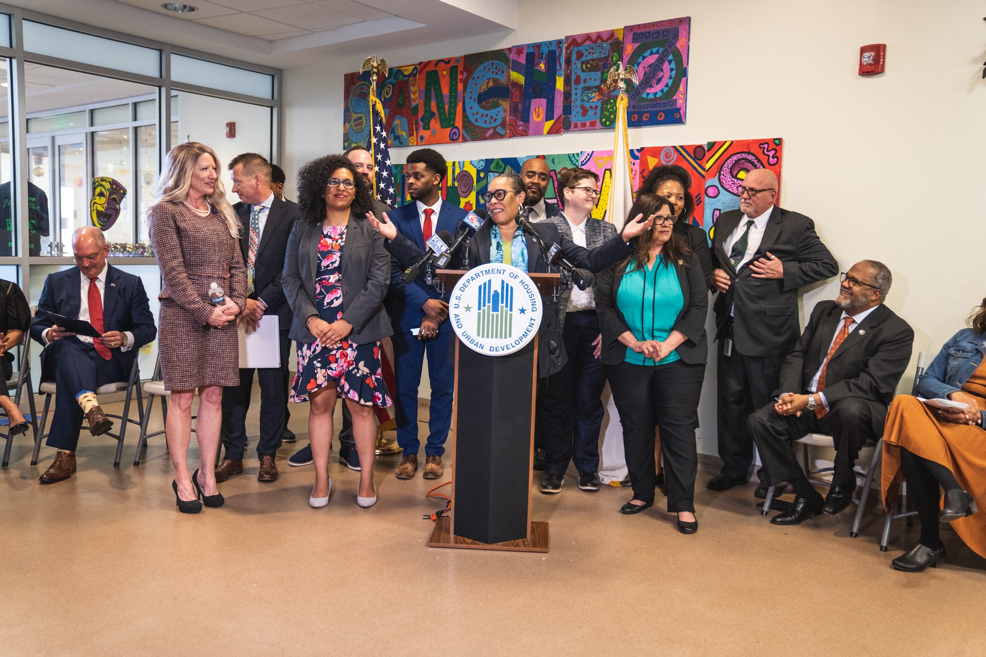 Gruppe von Menschen vor einem Rednerpult mit Mikrofon und einer Tafel mit dem Text "Sancho-Stadtrat tritt dem Stadtrat bei", mit sitzenden und stehenden Teilnehmern, Flaggen, Feuerlöscher und Deckenleuchten im Hintergrund.