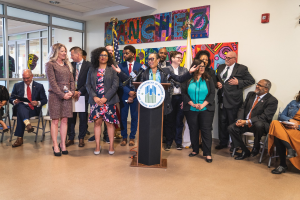 Gruppe von Menschen vor einem Rednerpult mit Mikrofon und einer Tafel mit dem Text "Sancho-Stadtrat tritt dem Stadtrat bei", mit sitzenden und stehenden Teilnehmern, Flaggen, Feuerlöscher und Deckenleuchten im Hintergrund.