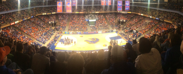 Eine große Halle mit Zuschauern, die ein Basketballspiel auf dem Platz verfolgen, umgeben von stehenden Zuschauern, sitzenden Fans in den Rängen und Deckenleuchten und Bannern.