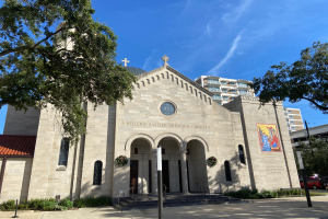 Die griechisch-orthodoxe Kirche in San Antonio, Texas, ist ein großer Bau mit Fenstern, Säulen, Bogengängen, Treppen mit Geländern und ist von Pflanzen, Bäumen und Fahrzeugen auf der Straße umgeben, unter einem bewölktem Himmel.